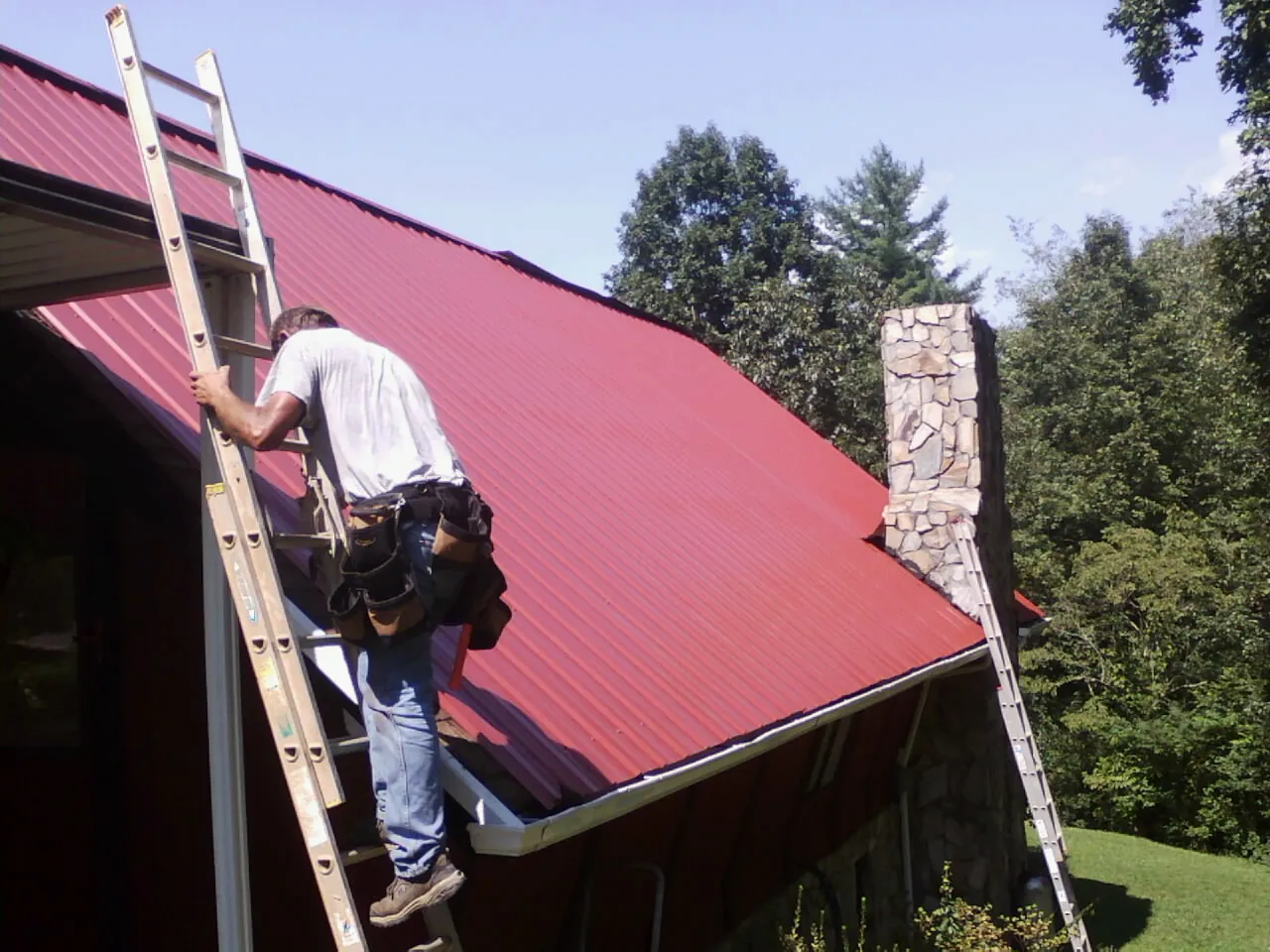 Roofer on a ladder beside a red metal roof and stone chimney — Soffit & Fascia Repair by Roofing Brothers in Stone Mountain